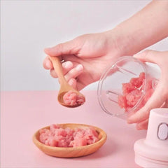 Person using a baby food processor to make pink baby food, with a wooden spoon and bowl on a pink surface.