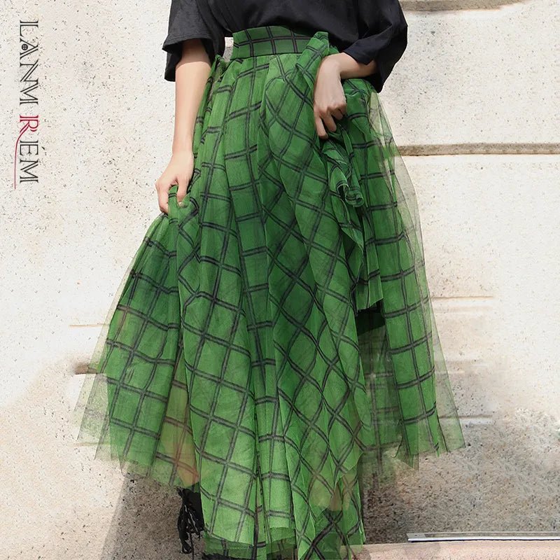 A woman wearing a green plaid tulle skirt with an asymmetric elastic waist and a high-waist silhouette. The skirt reaches mid-calf and is paired with a black top and black boots.