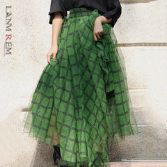 A woman wearing a green plaid tulle skirt with an asymmetric elastic waist and a high-waist silhouette. The skirt reaches mid-calf and is paired with a black top and black boots.