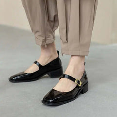 Black patent leather shoes with gold buckles worn with beige pants on a neutral background