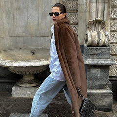 Woman in a brown coat and sunglasses walking past classical architecture.