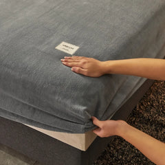Person adjusting a fitted sheet on a bed with a visible brand label.
