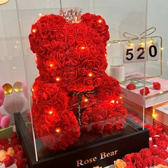 Heart-shaped rose bear with lights inside a glass display case, surrounded by more roses and a calendar. red