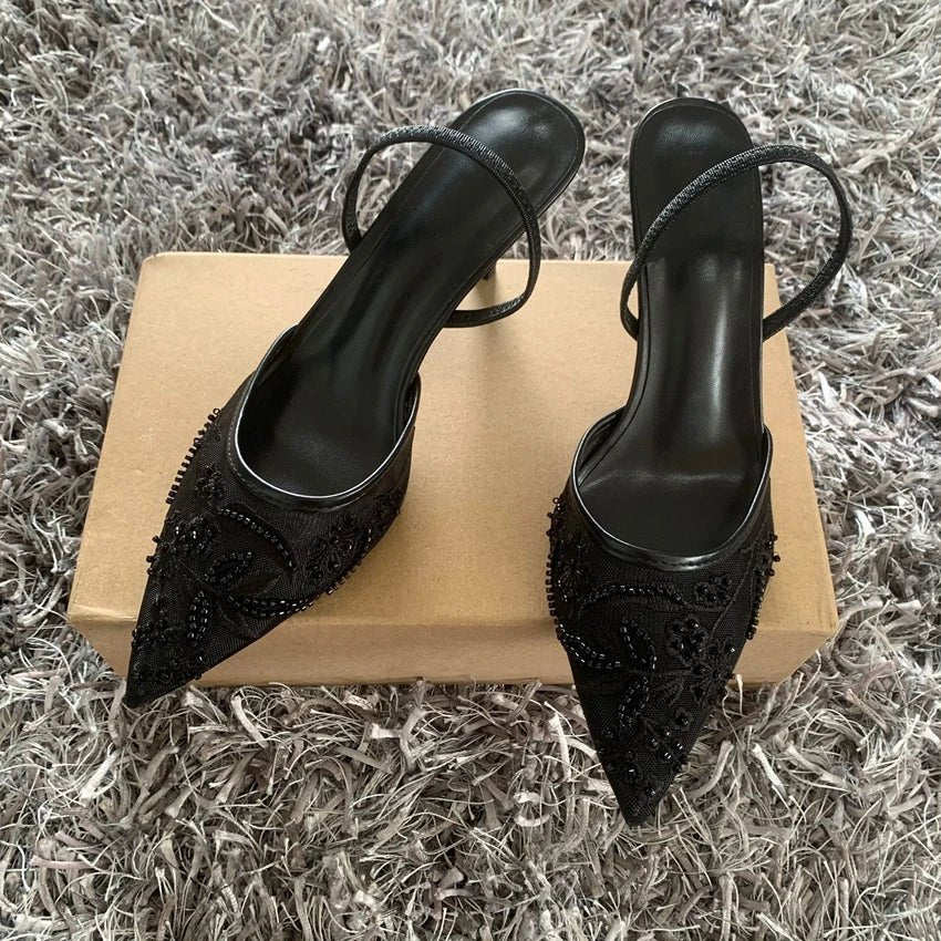 Black high-heeled shoes with embellishments on a cardboard box against a textured gray background