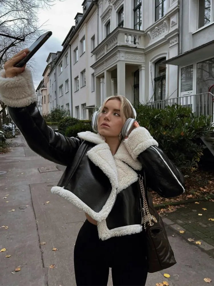 Woman wearing a black faux leather jacket with thick white fur lapels, paired with a black hat and earmuffs, taking a picture with a smartphone.