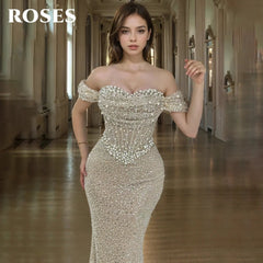 Woman in a sparkling off-shoulder gown standing in an elegant hallway with 'ROSES' branding.