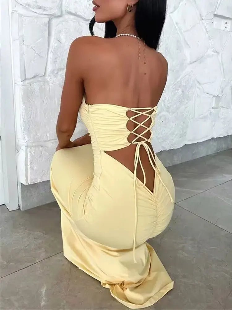 A woman wearing a strapless, off-shoulder, yellow maxi dress with a lace-up back detail, standing in a kneeling position with her back towards the camera.