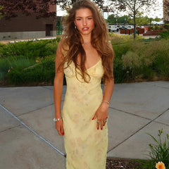 Woman in a yellow dress standing outdoors on a sidewalk with greenery and buildings in the background.