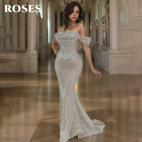 Woman in a sparkling off-shoulder gown standing in an elegant hallway with 'ROSES' branding.