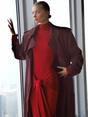 Woman in a red dress and dark coat standing by a window with a cityscape view.