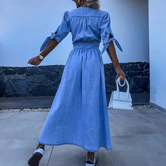 Woman wearing a blue dress holding a white handbag in an outdoor setting.