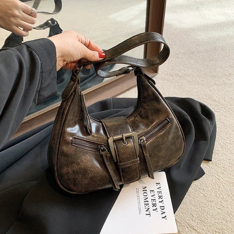 Brown leather handbag with a strap held by a person, placed on a surface with a magazine underneath.