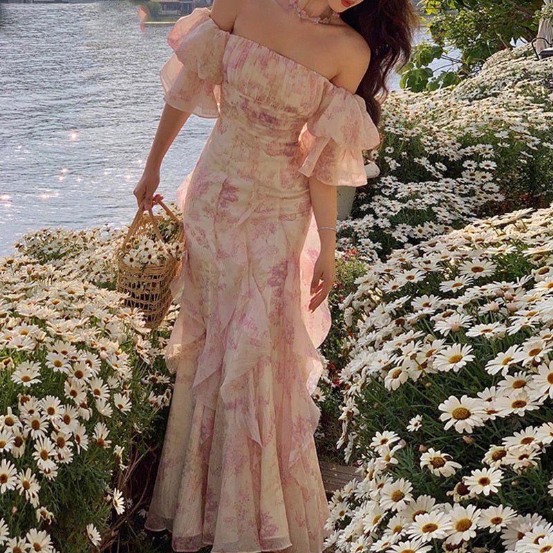 A woman wearing a midi-length, floral chiffon dress with spaghetti straps and a fairy-core aesthetic, standing by a flower bed.