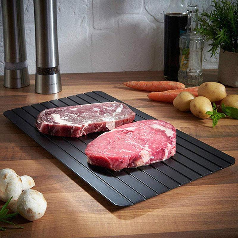 A black, rectangular defrosting tray with thawed meat on it, placed on a wooden surface with vegetables in the background.