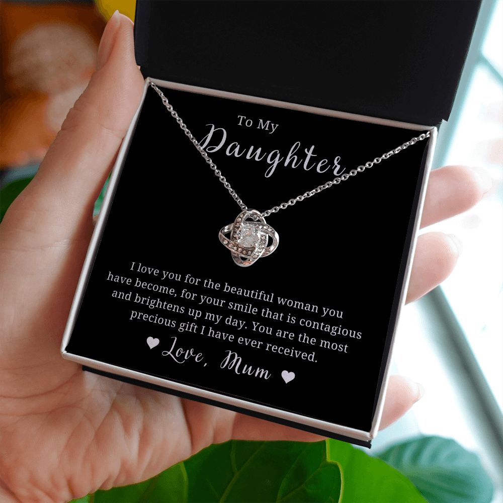 A necklace with a silver or gold colored pendant in the shape of a love knot, displayed in a black box with a message card.