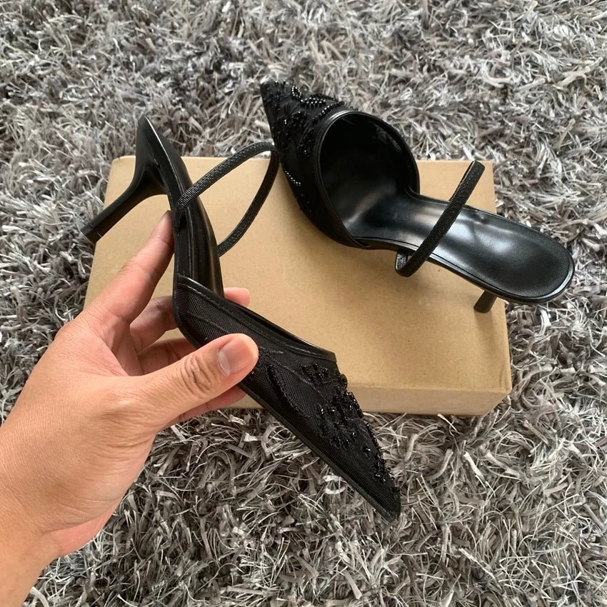 Black high-heeled shoes with intricate designs held by a hand on a textured gray surface.