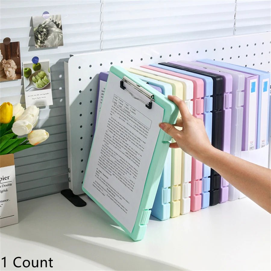 Colorful file folders on a desk with a hand holding one, next to a pegboard with photos and a vase of flowers.