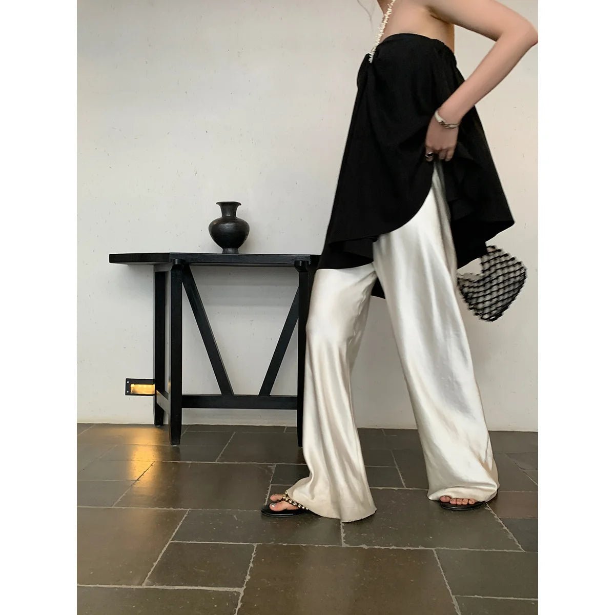 Person wearing a black top and white pants standing in a room with a black table and vase.