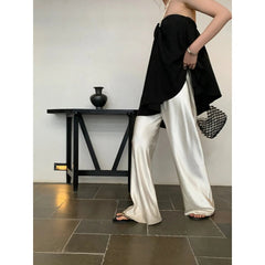 Person wearing a black top and white pants standing in a room with a black table and vase.