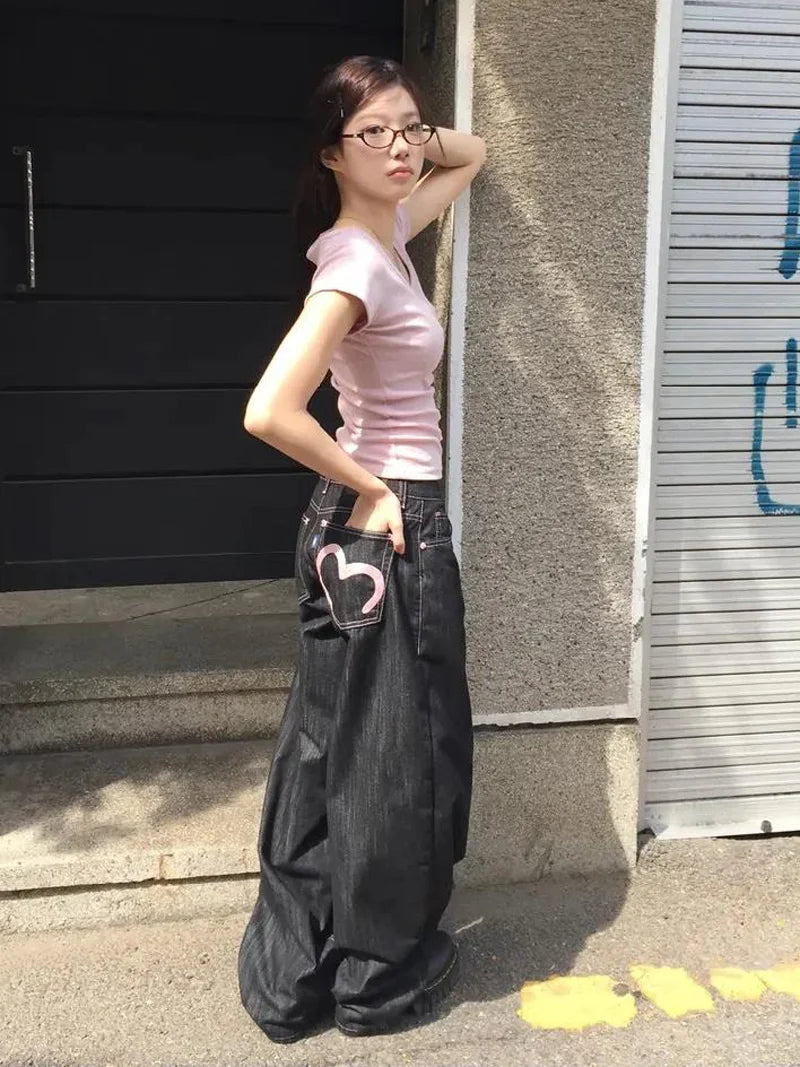 Woman wearing a pink top and black wide-leg pants leaning against a wall.