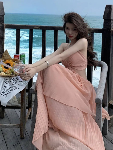 Woman in a peach dress sitting on a wooden deck by the ocean, holding a glass.