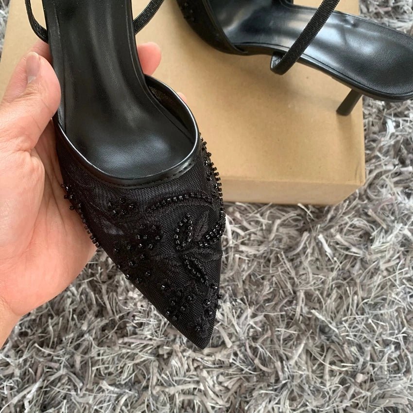 Black high-heeled shoe with intricate design held by a hand on a textured surface.