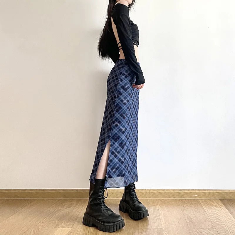 Person wearing a black long-sleeve top and blue plaid skirt with a high slit, standing against a plain wall.