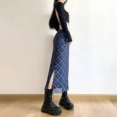 Person wearing a black long-sleeve top and blue plaid skirt with a high slit, standing against a plain wall.