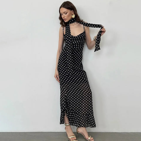A woman models a black halter-neck maxi dress with a white polka dot pattern. She is holding the tie at the back of her neck and wearing gold earrings and heeled sandals against a plain white wall.
