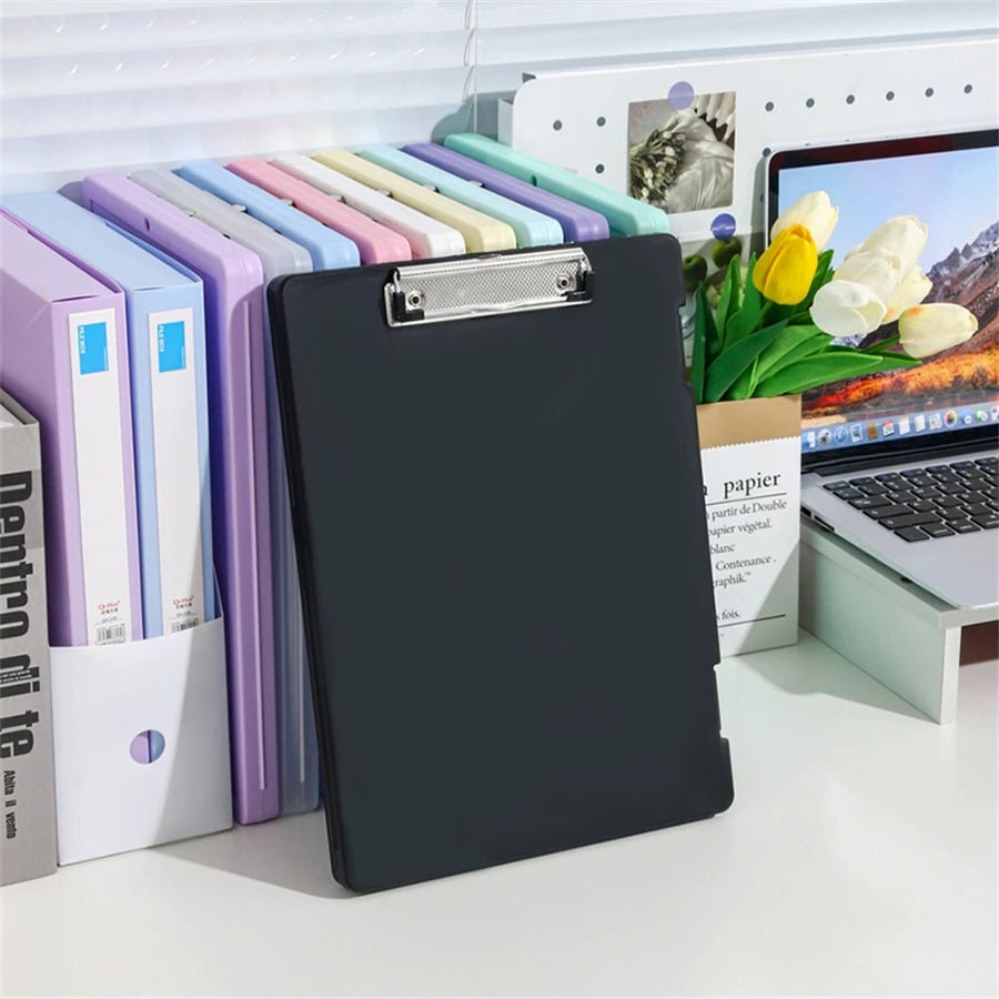 Black clipboard on a desk with office supplies including colorful folders and a laptop.