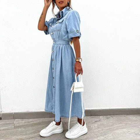 Woman wearing a denim dress with white sneakers and a small white handbag on a plain background