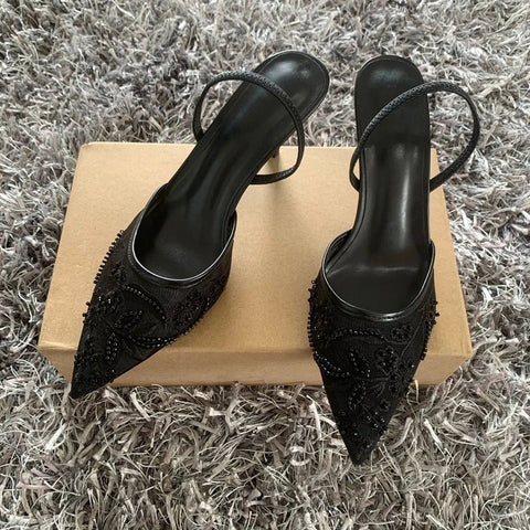 Black high-heeled shoes with embellishments on a cardboard box against a textured gray background