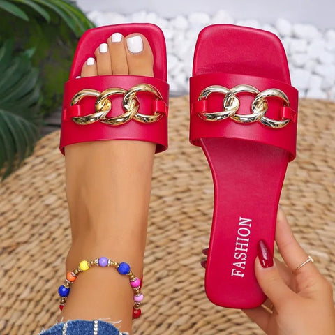 Red sandals with gold chain detail worn by a person on a woven surface.