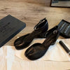 Pair of black high-heeled shoes on a wooden floor with a box and other items.