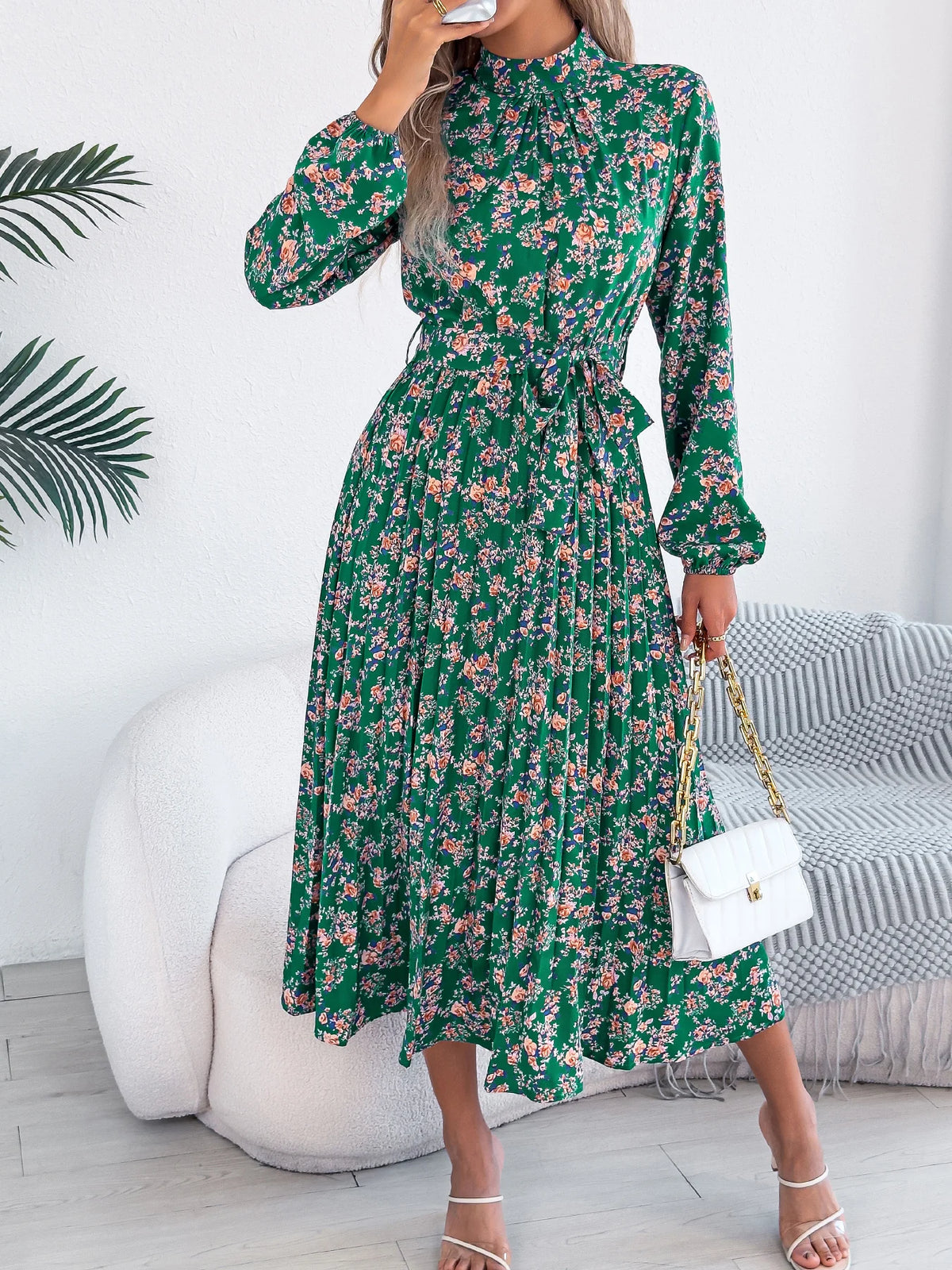 Woman wearing a green floral dress in a room with a white wall and gray sofa.