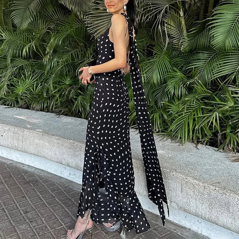Woman in a black polka dot dress with a long train standing outdoors against green foliage.