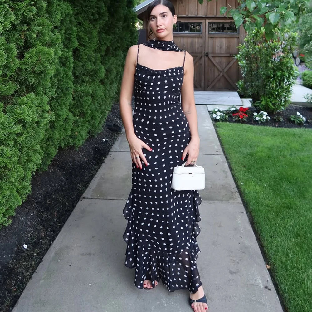 Woman in a black polka dot dress standing on a sidewalk with greenery in the background