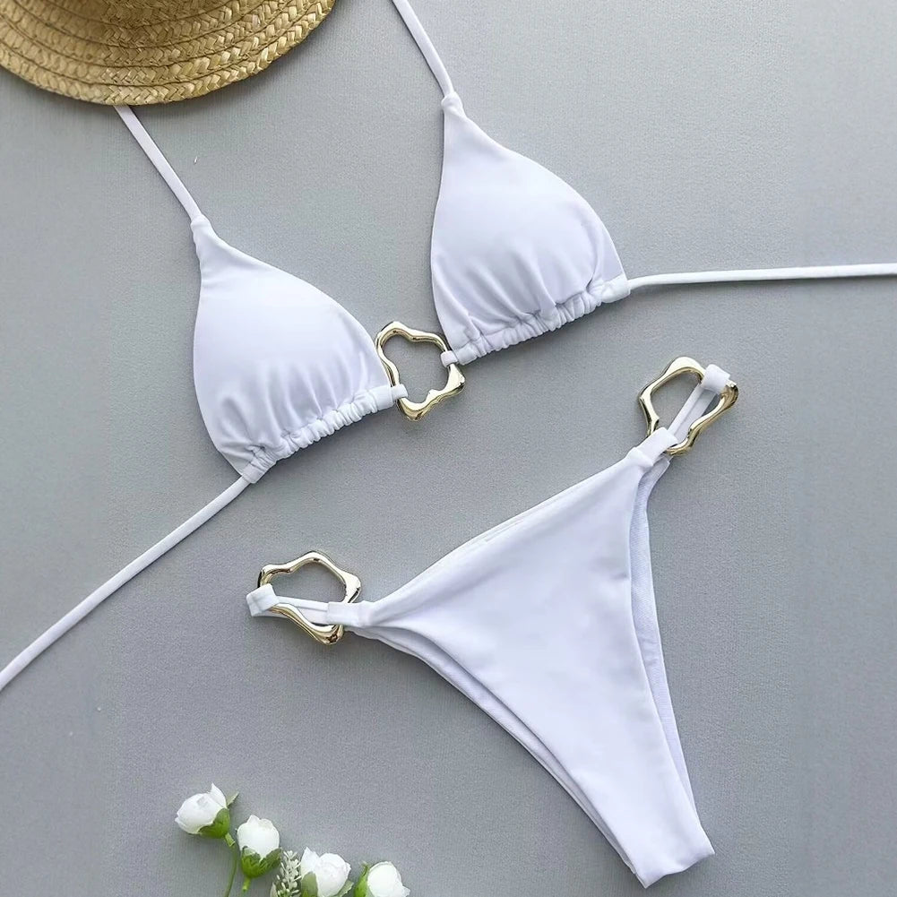 White bikini set with gold accents on a gray surface, accompanied by a straw hat and flowers.