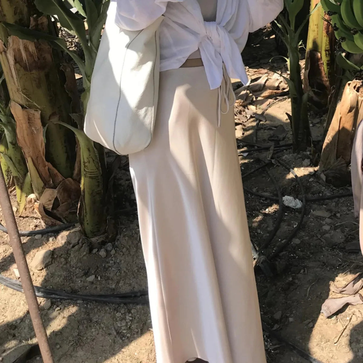 Person wearing a white top and beige pants standing in a natural setting with plants.