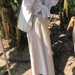 Person wearing a white top and beige pants standing in a natural setting with plants.