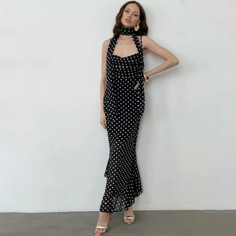 A woman models a black halter-neck maxi dress with a white polka dot pattern, standing against a plain white wall.