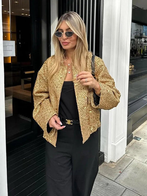 Woman wearing a gold jacket and black pants standing outside a building.