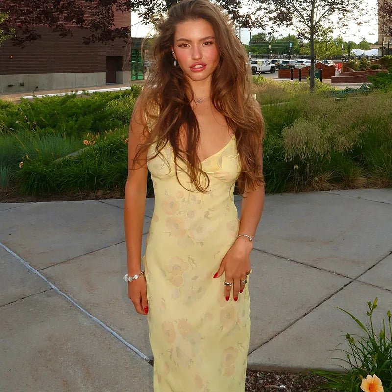 Woman in a yellow dress standing outdoors on a sidewalk with greenery and buildings in the background.