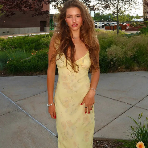 Woman in a yellow dress standing outdoors on a sidewalk with greenery and buildings in the background.