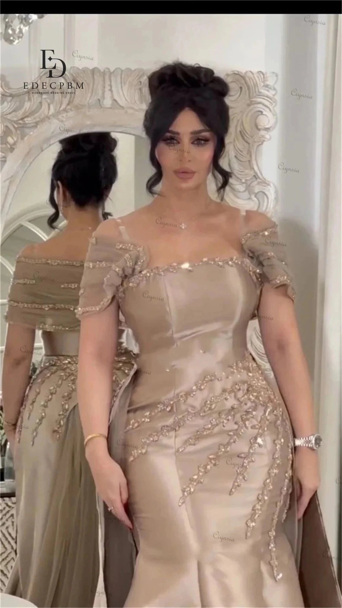 Woman in a beige evening gown with intricate detailing standing in front of a decorative mirror.