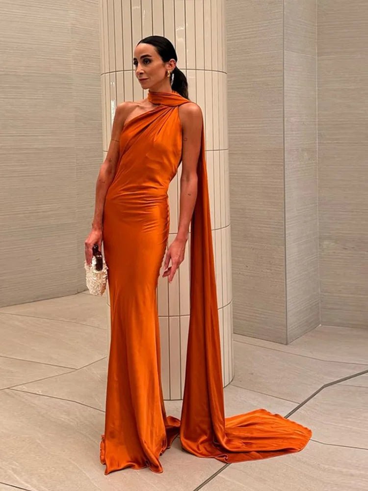 Woman in an orange evening gown with a long train standing against a neutral wall.