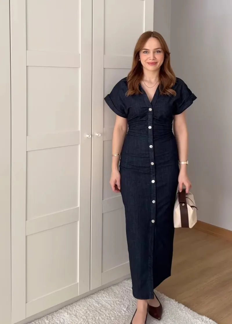Woman in a dark blue dress standing in a room with light-colored walls and wooden floor.