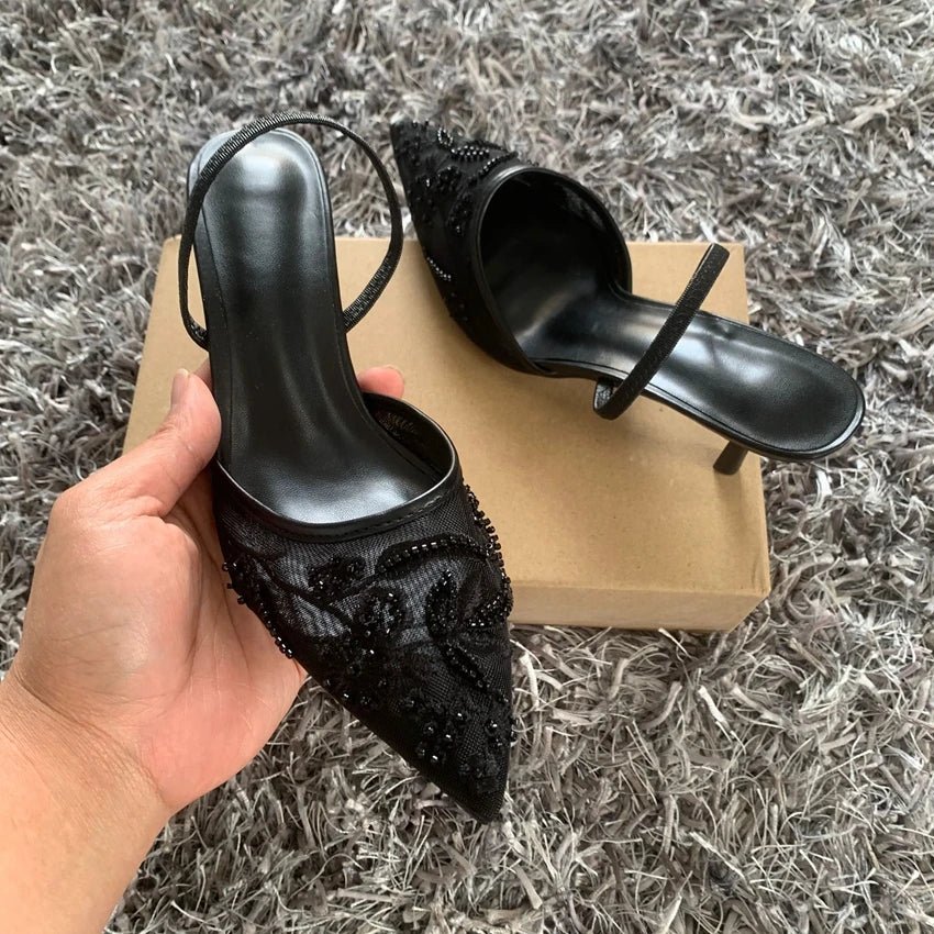 Black high-heeled shoes with embellishments held by a hand on a textured gray surface.