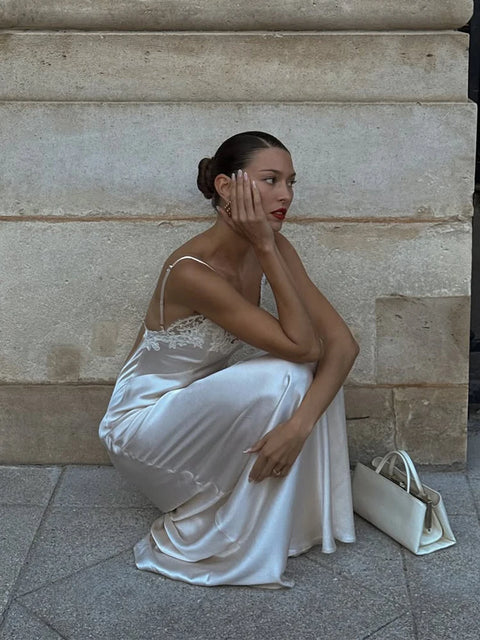 Woman in a white dress sitting against a stone wall with a handbag beside her.