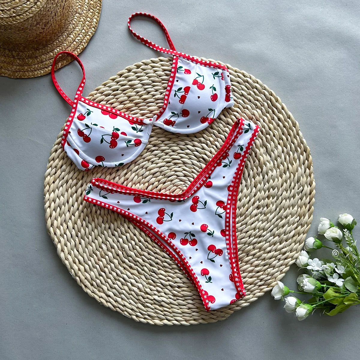 White bikini with red cherries on a woven mat with a straw hat and flowers.